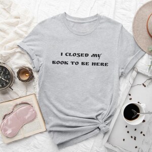 May include: A light gray t-shirt with the text "I CLOSED MY BOOK TO BE HERE" in black. The shirt is knotted at the waist. A book, eye mask, coffee, and a candle are next to the shirt.