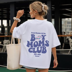 May include: White t-shirt with the text "Proud Member Of The Boy Moms Club" in blue, featuring a car graphic. The shirt also includes "Est. 2026" and the name "Manuel".