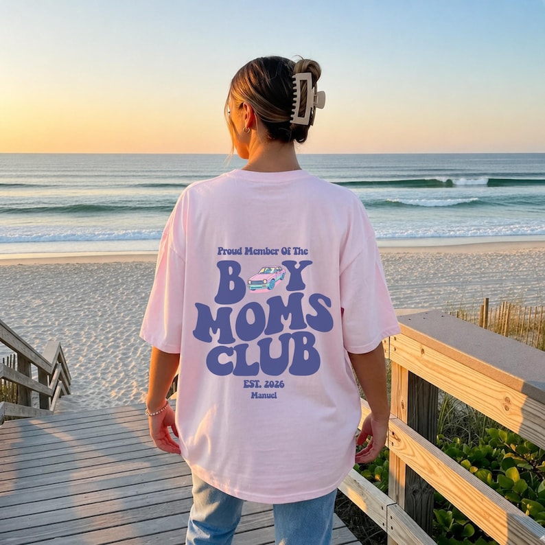 May include: A light pink t-shirt with the text "Proud Member Of The Boy Moms Club" in blue. The design includes a car graphic and the text "Est. 2026 Manual". The shirt is worn on a beach.