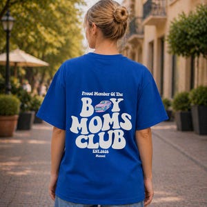 May include: A royal blue t-shirt with white text that reads "Proud Member Of The Boy Moms Club." The graphic includes a pink and blue car illustration. The shirt also has the text "EST.2026 Manuel."