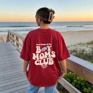 May include: A red t-shirt with the text "Proud Member Of The Boy Moms Club" in a retro font. The design includes a light blue car graphic and the text "EST.2026". The shirt is worn by a person on a wooden boardwalk near the beach.