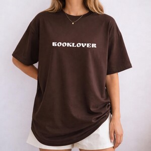 May include: A brown t-shirt with the word "BOOKLOVER" printed in white across the chest. The shirt has short sleeves and a relaxed fit. The model is wearing white shorts.