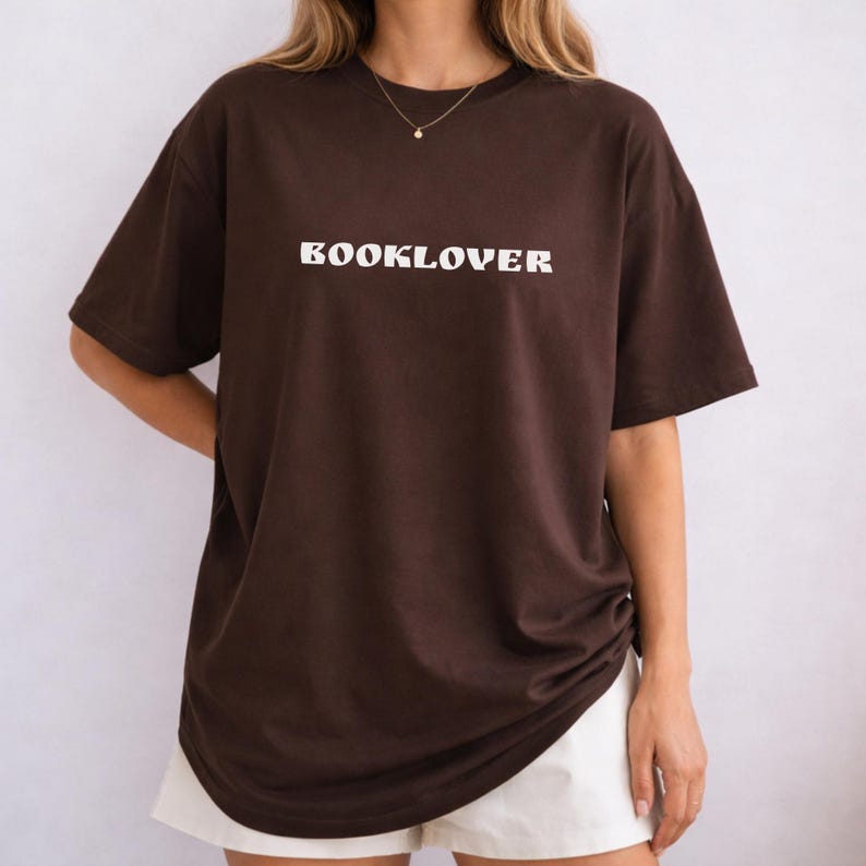 May include: A brown t-shirt with the word "BOOKLOVER" printed in white across the chest. The shirt has short sleeves and a relaxed fit. The model is wearing white shorts.