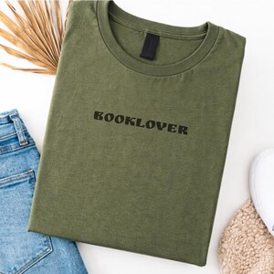 May include: An olive green t-shirt with the word "BOOKLOVER" printed in black. The shirt is folded and placed on a white surface, with a pair of blue jeans, a dried palm leaf, and a white shoe visible.