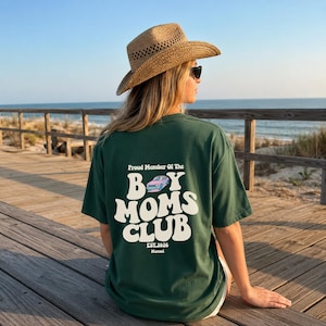 May include: Dark green t-shirt with the text "Proud Member Of The Boy Moms Club" in white, along with a car graphic. The shirt also includes "EST.2026" and the name "Manuel". The person is wearing a straw hat and sunglasses.