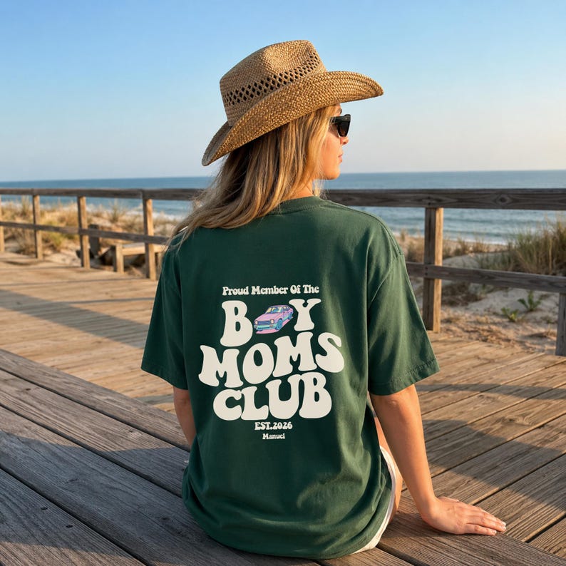 May include: Dark green t-shirt with the text "Proud Member Of The Boy Moms Club" in white, along with a car graphic. The shirt also includes "EST.2026" and the name "Manuel". The person is wearing a straw hat and sunglasses.