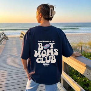 May include: Navy blue oversized t-shirt with the text "Proud Member Of The Boy Moms Club" in white, along with a car graphic. The shirt also includes "EST. 2026" and "Manual" in white. The image is taken on a wooden boardwalk overlooking the beach.