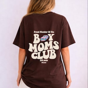 May include: A brown t-shirt with the text "Proud Member Of The Boy Moms Club" in white, along with a graphic of a car. The shirt also includes "EST.2026 Manuel". The person is wearing white shorts.