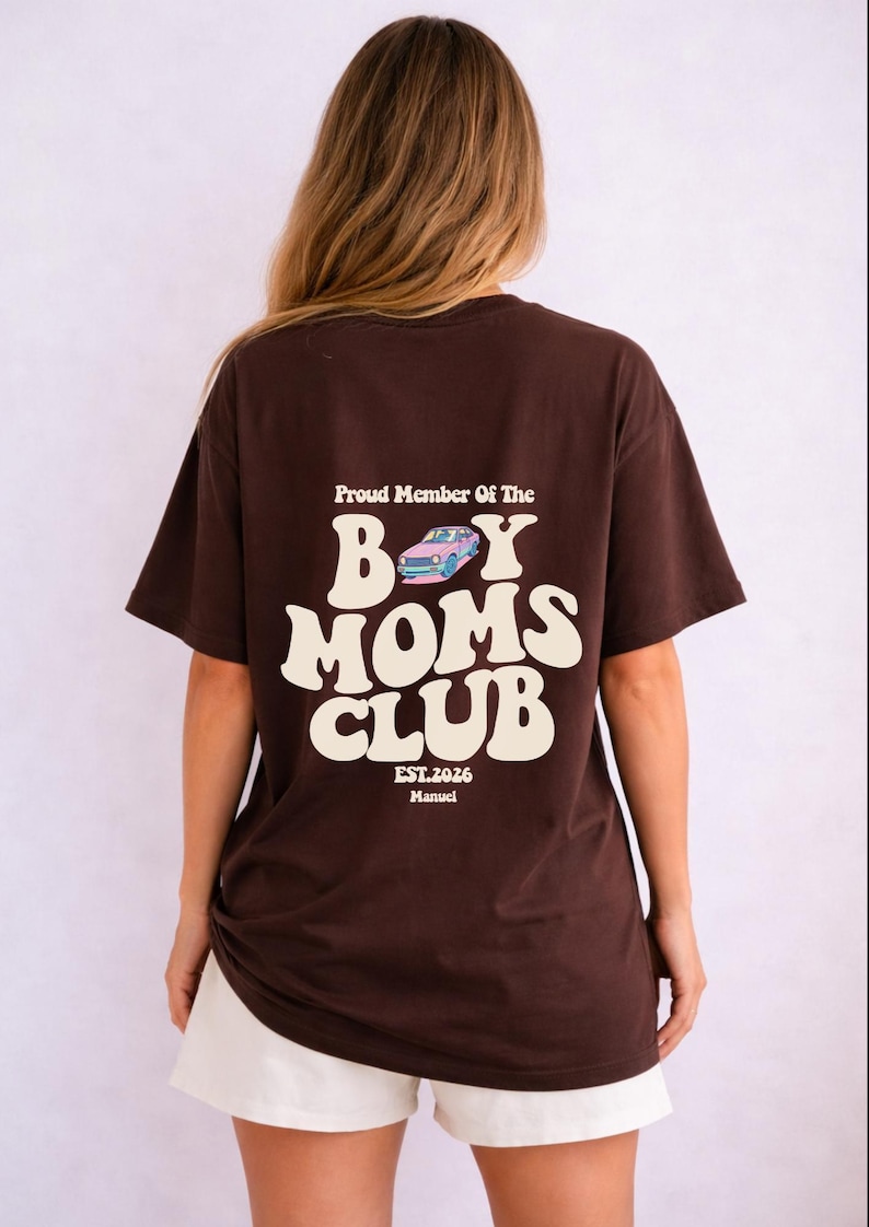 May include: A brown t-shirt with the text "Proud Member Of The Boy Moms Club" in white, along with a graphic of a car. The shirt also includes "EST.2026 Manuel". The person is wearing white shorts.
