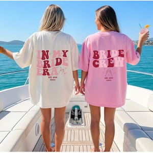 Könnte beinhalten: Zwei Frauen tragen übergroße T-Shirts mit Brautparty-Designs. Eines ist cremefarben mit dem Text "IN MY BRIDE ERA" in Rosa und Rot. Das andere ist rosa mit "BRIDE CREW" in ähnlichen Farben. Beide Shirts haben zusätzlichen Text und Grafiken.