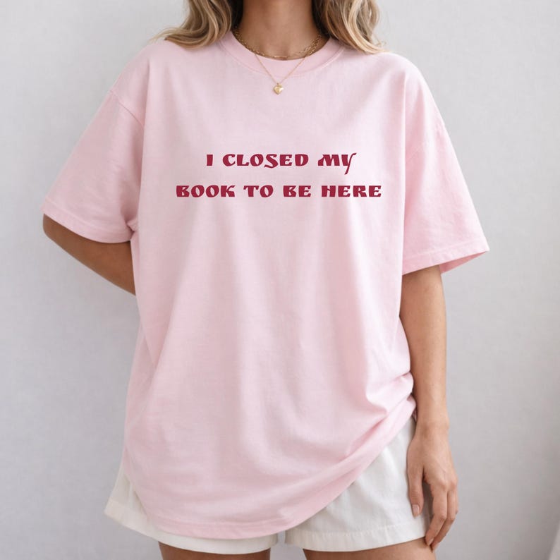 May include: A light pink t-shirt with the text "I CLOSED MY BOOK TO BE HERE" in burgundy lettering. The shirt has short sleeves and a relaxed fit.