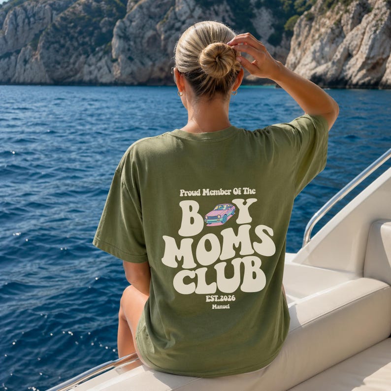 May include: Olive green t-shirt with the text "Proud Member Of The Boy Moms Club" in a retro font. The design includes a pink and blue car graphic. The shirt also has "EST.2026" and "Manual" printed below the main text.