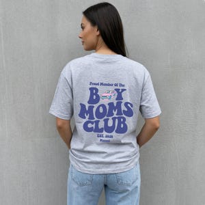 May include: A gray t-shirt with the text "Proud Member Of The Boy Moms Club" in blue, with a car graphic. The shirt also has the text "Est. 2026 Manuel" in a smaller font. The shirt is being worn.