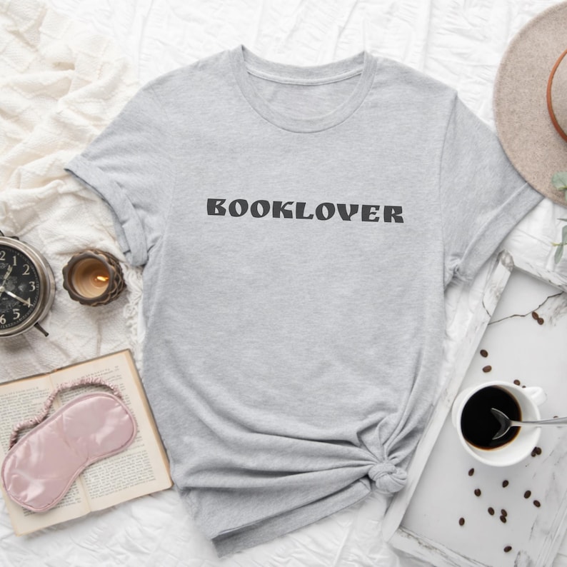 May include: A light gray t-shirt with the word "BOOKLOVER" printed in black. The shirt is styled with a book, eye mask, coffee, and a candle. The shirt is knotted at the waist.