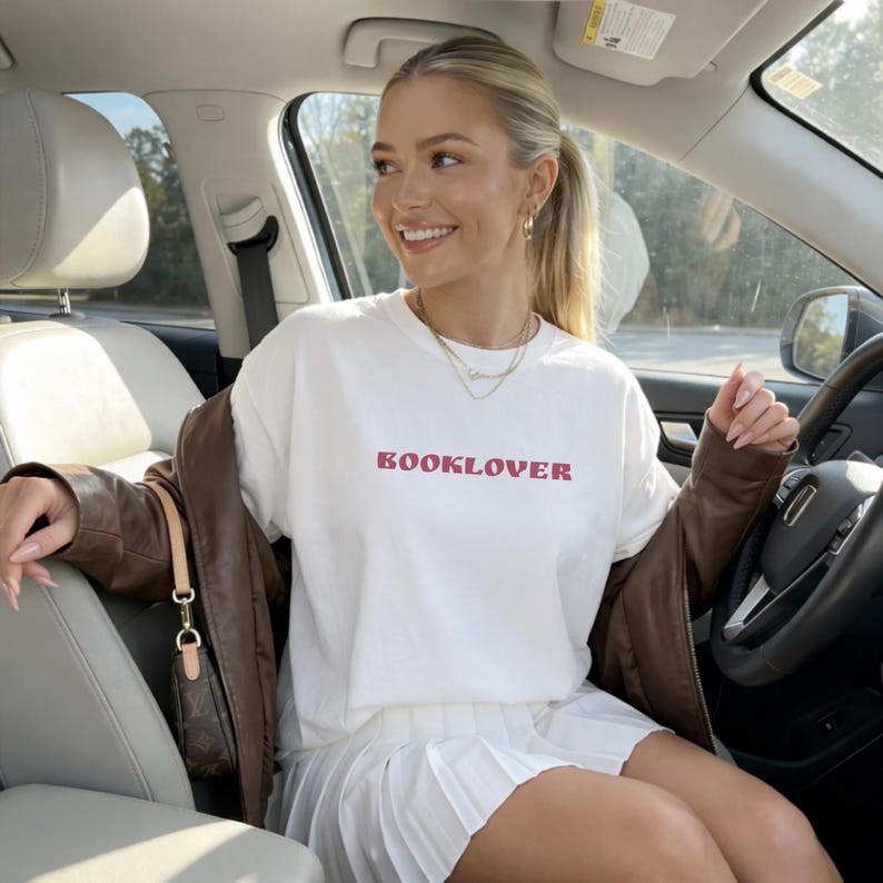 May include: White t-shirt with the word "BOOKLOVER" in red lettering. The person is wearing a brown leather jacket and a white pleated skirt. The person is sitting in a car.