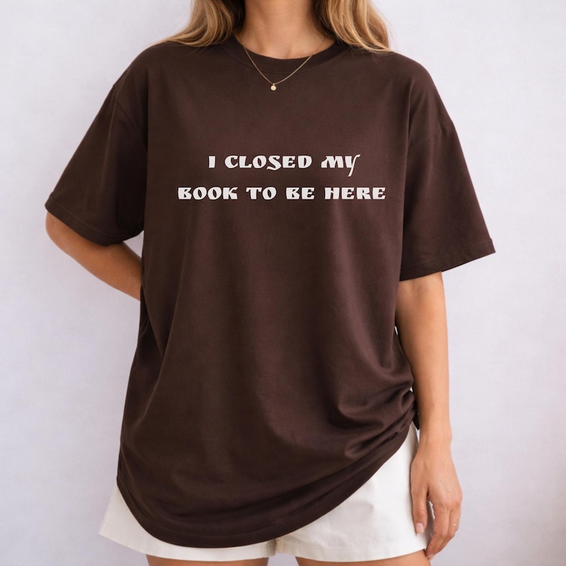 May include: A brown t-shirt with the text "I CLOSED MY BOOK TO BE HERE" in white, centered on the front. The shirt is a loose fit and appears to be made of a soft material. The model is wearing white shorts.