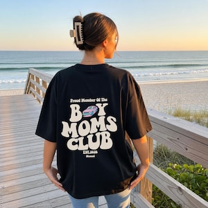 May include: Black t-shirt with the text "Proud Member Of The Boy Moms Club" in a retro font. The design includes a light blue car graphic and the text "EST.2026". The shirt is worn by a person on a wooden walkway overlooking the ocean.
