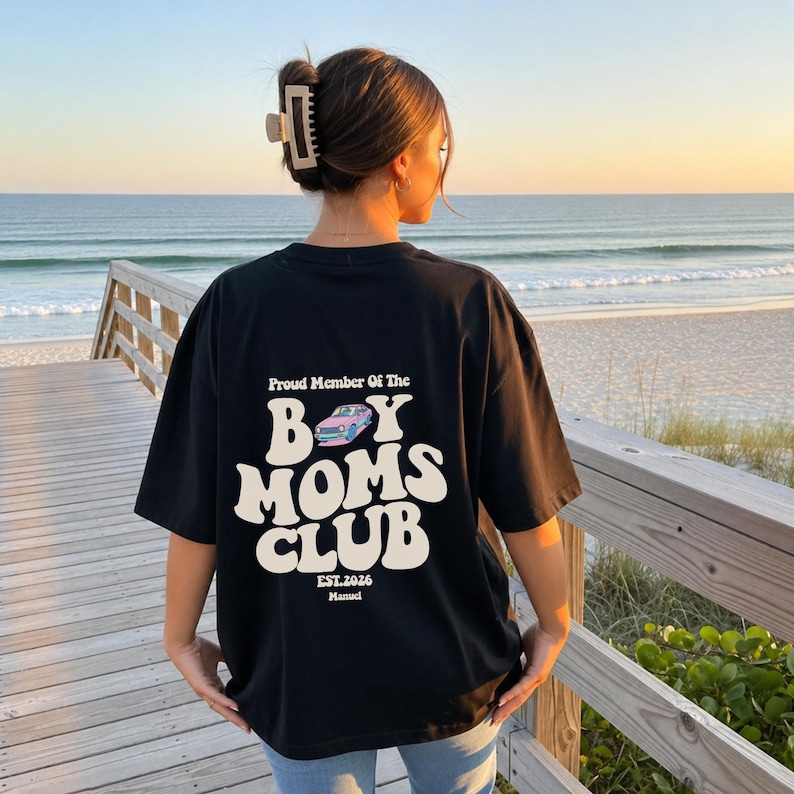May include: Black t-shirt with the text "Proud Member Of The Boy Moms Club" in a retro font. The design includes a light blue car graphic and the text "EST.2026". The shirt is worn by a person on a wooden walkway overlooking the ocean.