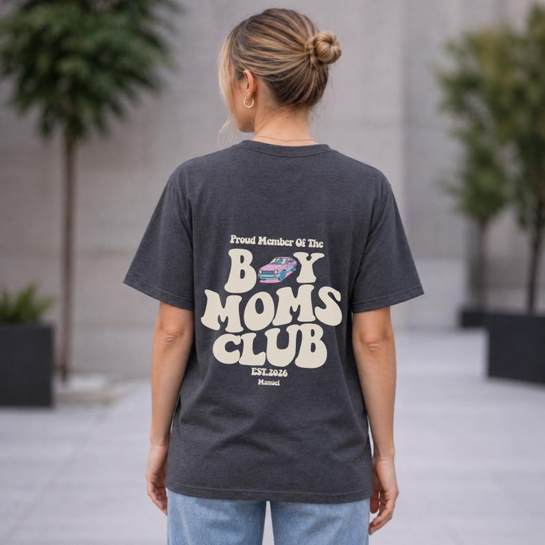 May include: Dark gray t-shirt with the text "Proud Member Of The Boy Moms Club" in a retro font. The design includes a pink and blue car graphic. The shirt also has "EST.2026" and "Manual" printed below the main text.