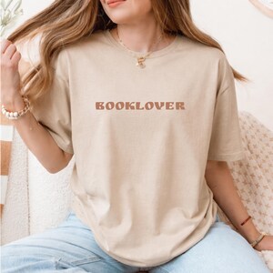 May include: A beige t-shirt with the word "BOOKLOVER" printed in a brown, retro font across the chest. The shirt has short sleeves and a relaxed fit. The model is wearing light blue jeans.