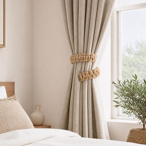 May include: A pair of beige, textured fabric curtains held back by two thick, hand-knotted natural jute rope tiebacks. The curtains feature a vertical weave pattern and are draped elegantly in front of a window, creating a soft, neutral aesthetic.
