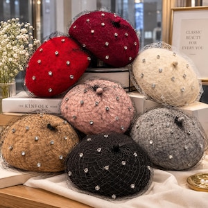 May include: A collection of six wool berets in burgundy, red, cream, pink, tan, and black. Each beret features a black mesh veil overlay and is embellished with scattered clear rhinestones across the surface of the fabric.