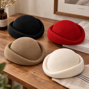May include: Four dome-shaped hats with rolled brims, presented in solid black, vibrant red, neutral beige, and off-white colors. Each hat features a smooth, felt-like texture and a rounded crown design, arranged together on a wooden surface.