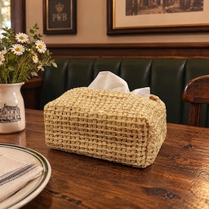May include: A rectangular tissue box cover crafted from woven light tan natural fibres. The box features an open top design with white tissues protruding from the centre. The woven texture creates a grid-like pattern across the exterior surfaces.
