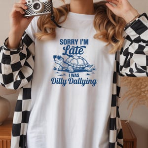 May include: A white long-sleeve shirt with black and white checkered sleeves. The front features a blue graphic of a turtle wearing heart-shaped sunglasses, with the text SORRY I'M Late I WAS Dilly Dallying printed in blue above and below the turtle.