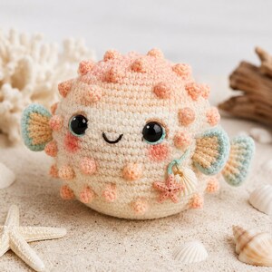 May include: A round, cream-colored crochet pufferfish toy with soft peach-colored spikes. It features large black eyes, a small embroidered smile, pink cheeks, and light blue fins. A small charm with a starfish and seashell hangs from its side.