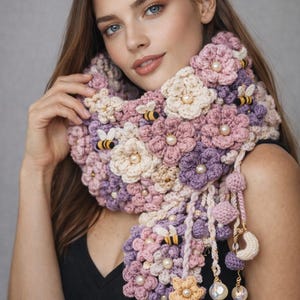 May include: A handmade crochet scarf featuring a floral design in shades of pink, purple, and cream. The scarf is adorned with pearl accents and small bee appliqués. Tassels and decorative charms hang from the ends.