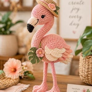 May include: A crocheted pink flamingo figurine wearing a tan straw hat adorned with a small pink flower and green leaves. The flamingo has a black-tipped beak, a light pink wing, and holds a green monstera leaf with a pink flower in its wing.