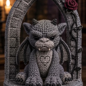 Pode incluir: Uma figura de gárgula em crochê cinza com pequenos chifres, asas e dois dentes brancos, sentada dentro de um arco de crochê que imita pedra. O arco apresenta uma rosa vermelha escura em crochê e um padrão de videira, com um coração no peito.