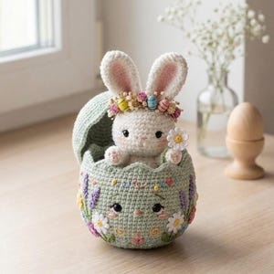Puede incluir: Un conejito de Pascua de ganchillo con una corona de flores, saliendo de una cáscara de huevo verde pastel. La cáscara está decorada con flores y hojas bordadas. Una encantadora decoración primaveral.