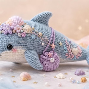 May include: A crocheted dolphin in shades of blue, white, and pink, adorned with a seashell crown, floral accents, and a small purple purse. The dolphin has a smiling expression and is decorated with embroidered details.
