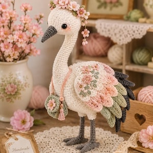 May include: A handcrafted crochet stork figurine, adorned with a floral crown and decorative wings in shades of pink, green, and gray. The stork carries a small purse and stands on a crocheted doily. A tag reads "Handmade with love."