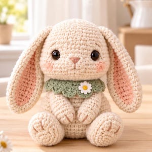 May include: A cream-colored crochet rabbit plush toy with long ears featuring pink inner sections. The rabbit has large dark eyes, pink cheeks, and wears a sage green scalloped collar adorned with a small white daisy flower applique.