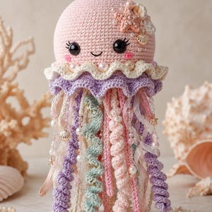 May include: A crocheted jellyfish toy in pastel pink, featuring a rounded head with black safety eyes, a smiling mouth, and pink cheeks. It is adorned with a cream and lavender ruffled skirt, pearl beads, and dangling spiral tentacles in pink, purple, and mint green.