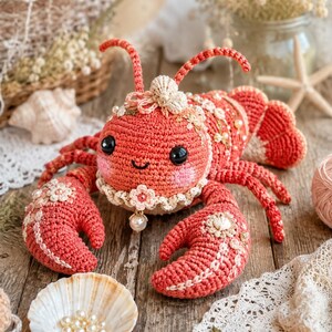 Puede incluir: Un juguete de langosta tejido a ganchillo en hilo color rojo coral, con grandes ojos negros de seguridad, dos antenas y dos pinzas grandes. La langosta está decorada con pequeñas flores blancas tejidas, perlas diminutas y una concha.