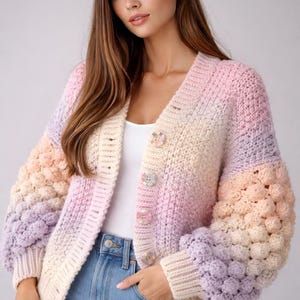 May include: A pastel-coloured, open-front cardigan with a V-neckline. The sweater features a gradient of colours, including pink, peach, lavender, and cream. The sleeves have a textured, bobble-stitch design. The cardigan is buttoned with decorative buttons.