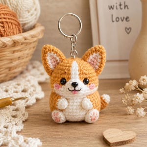 May include: A crochet keychain shaped like a Corgi dog with tan and white yarn, black plastic eyes, a small black nose, and pink stitched cheeks. The figure has a metal split ring attached to the top and features small paws with pink details.