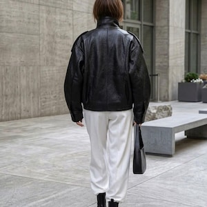 May include: A black leather jacket with a high collar and long sleeves, paired with white trousers, black ankle boots, and a black shoulder bag. The jacket features a textured finish and structured shoulders, creating a clean and cohesive look.