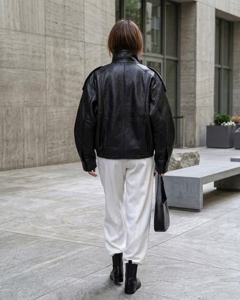 May include: A black leather jacket with a high collar and long sleeves, paired with white trousers, black ankle boots, and a black shoulder bag. The jacket features a textured finish and structured shoulders, creating a clean and cohesive look.
