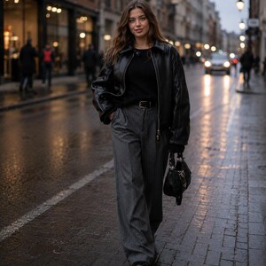 Peut inclure: Une veste en cuir noir portée sur un pull noir, associée à un pantalon large gris à fines rayures avec une ceinture noire. Un petit sac à main noir avec des détails argentés est porté, et des chaussures noires sont aux pieds.