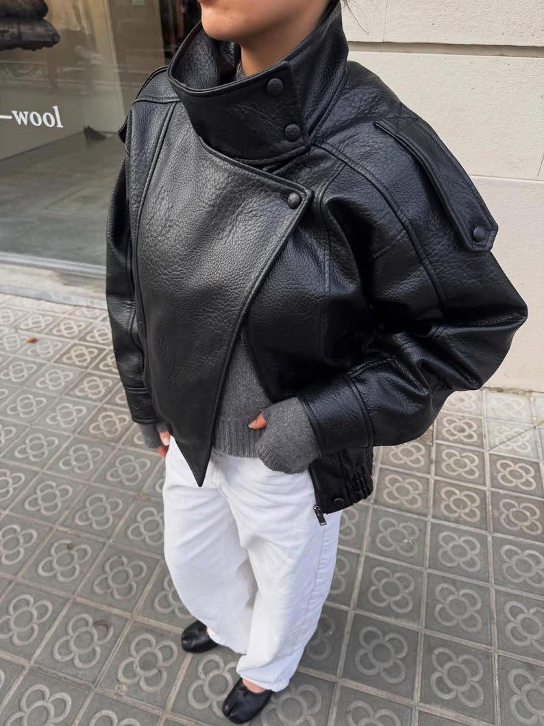 May include: A black leather jacket with a high collar, snap button closures, and shoulder epaulets. The jacket is worn over a grey sweater and white trousers, paired with black split-toe shoes. The leather has a textured, pebbled finish.