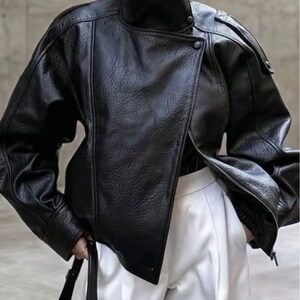 May include: A black leather jacket with a textured finish, featuring a high stand-up collar, an asymmetrical front closure with snap buttons, and long sleeves with dropped shoulders. The jacket is paired with white trousers.
