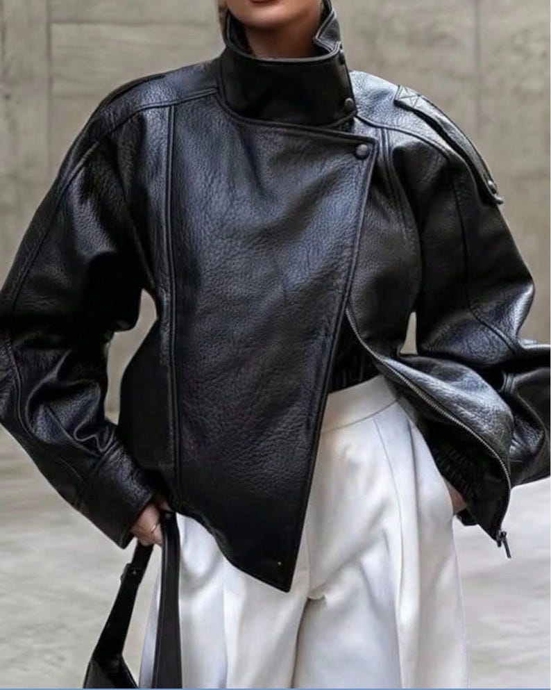 May include: A black leather jacket with a textured finish, featuring a high stand-up collar, an asymmetrical front closure with snap buttons, and long sleeves with dropped shoulders. The jacket is paired with white trousers.