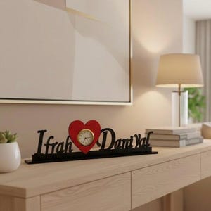 May include: A decorative black wooden sign with the text "Ifrah Danı Yal" and a red heart-shaped clock. A small succulent plant in a white pot sits to the left of the sign. The sign is on a light wood table.