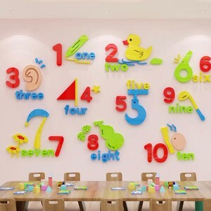 Puede incluir: Decoraciones de pared coloridas con los números del uno al diez, cada uno con su palabra correspondiente. Los números son de varios colores brillantes y diseños lúdicos, incluyendo un pato para el número dos. Una mesa con bloques y sillas en primer plano.