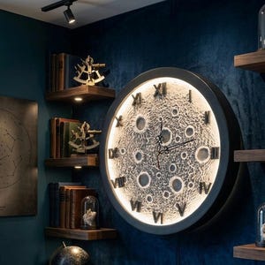 Puede incluir: Un gran reloj de pared redondo con una esfera lunar, con cráteres y números romanos iluminados. El reloj está colocado contra una pared azul oscuro, con estantes de madera que contienen libros y objetos decorativos. El diámetro del reloj es de aproximadamente 61 cm.